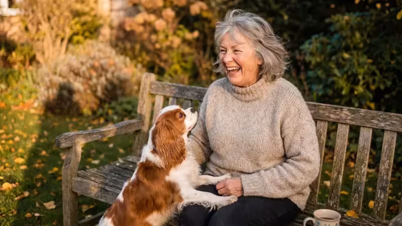 A smiling senior woman sits on a garden bench with a Cavalier King Charles Spaniel resting its paws on her lap in the autumn sun.
